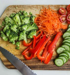 chopped broccoli carrots red pepper tomatoes and cucumber on a cutting board with a knife