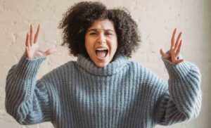 A woman with curly hair wearing a gray sweater screams with hands raised, expressing frustration.