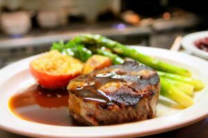 Succulent steak with a glossy, rich sauce, served with fresh asparagus, a roasted tomato topped with breadcrumbs, and golden potatoes on a white plate.