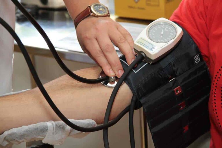 A blood pressure cuff around a person's upper arm, with a hand placing a stethoscope over the elbow joint.