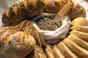 A variety of freshly baked breads, some topped with seeds, are artfully arranged around a bowl of mixed seeds with wheat stalks, creating a warm, rustic feel.