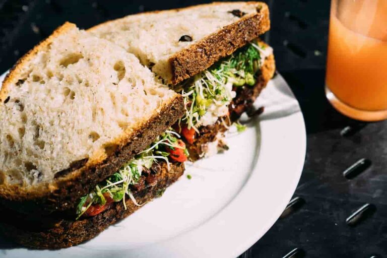 A close-up of a sandwich on a white plate, the sandwich filled with fresh greens, sprouts, and tomatoes, next to a glass of orange juice on a dark table.