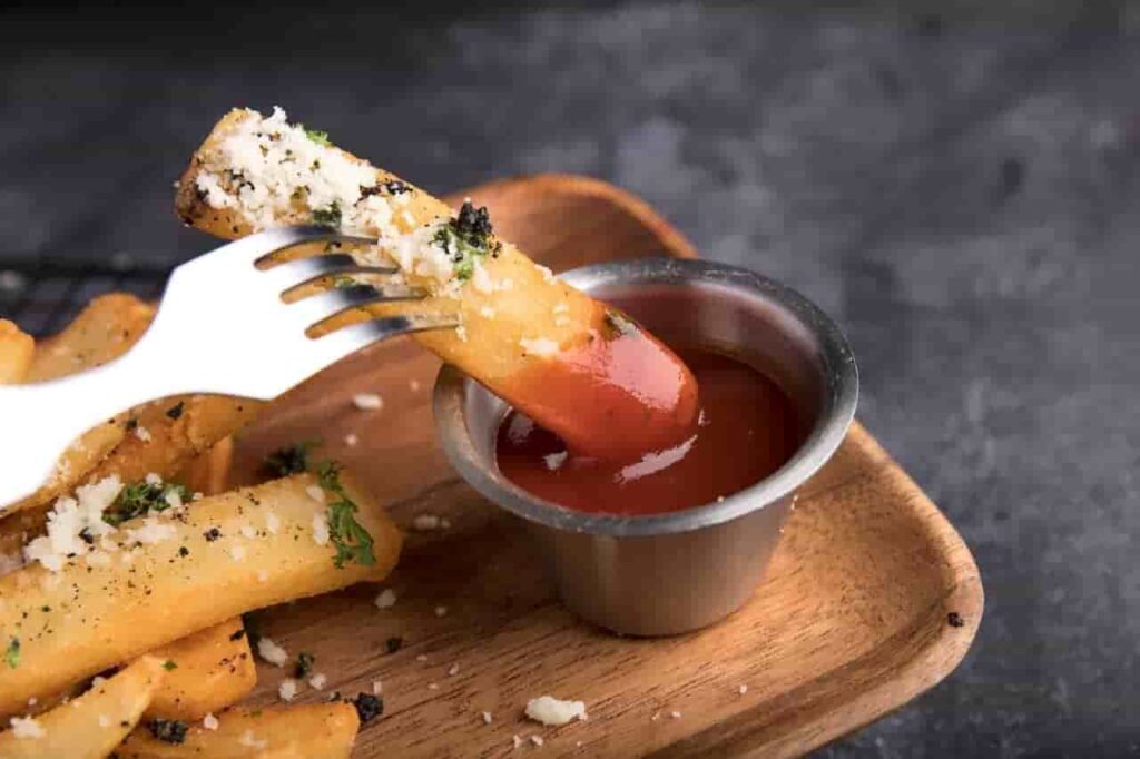 A fork dips a seasoned French fry with cheese and herbs into a silver cup of ketchup on a wooden tray, set against a dark background.