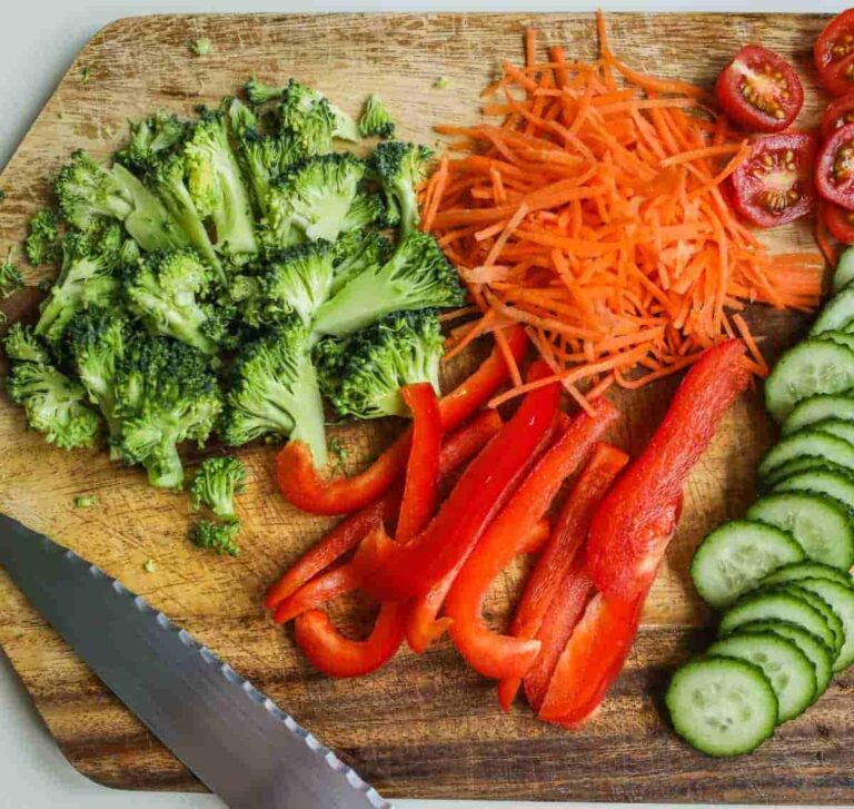 Chopped vegetables on a wooden board include broccoli florets, shredded carrots, sliced red bell peppers, halved cherry tomatoes, and cucumber slices, with a serrated knife nearby.