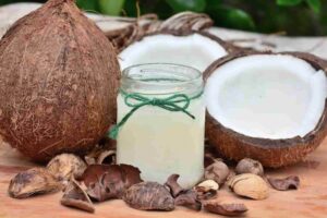 A jar of coconut oil with a green string sits on a table, surrounded by a whole coconut, cracked coconut halves, and scattered shells.