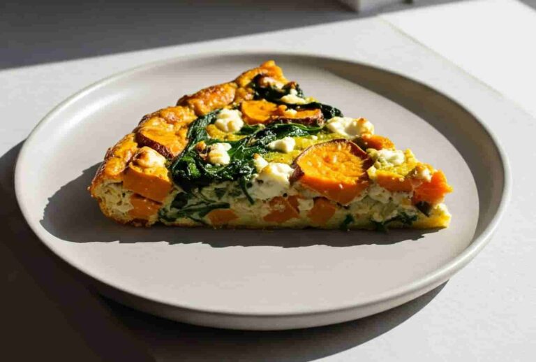 A slice of vegetable frittata on a white plate, featuring vibrant orange sweet potatoes, green spinach, and creamy white feta, illuminated by natural light.