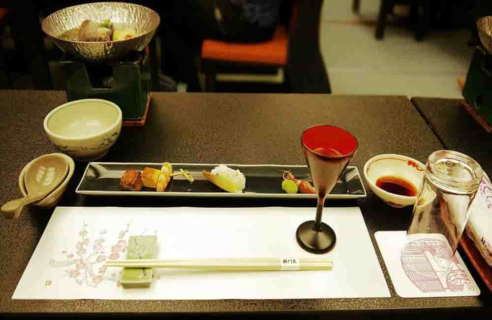 Elegant Japanese dining setup with a black platter of assorted appetizers, a red-stemmed glass, chopsticks on a floral mat, and bowls on a dark table.