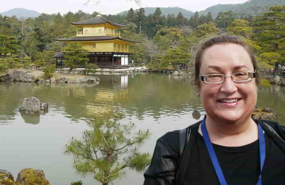 Natalia Knezevic smiles in the foreground, with the golden Kinkaku-ji temple and its serene reflection on a pond amidst lush greenery in the background.