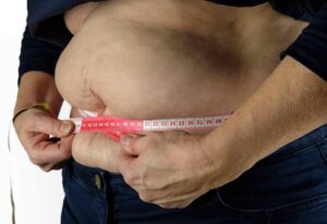 An obese person measuring their waist with a red measuring tape, pulling around their midsection slightly above the jeans, highlighting self-awareness and focus.