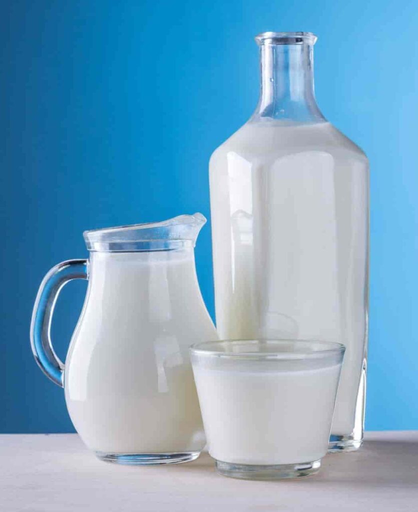 Three glass containers filled with milk are set against a smooth blue background. A tall bottle, a jug, and a glass create a clean, refreshing atmosphere.