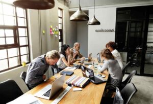 Six people in office casual clothing sitting around a long table in an open office area with a large window and glass doors to to another office area. Industrial light fixtures hang above the table. The people are working with laptops and notepads. There are post-it notes on a section of the wall behind the people.