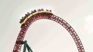 A yellow roller coaster car ascends an arching red track with visible riders excitedly raising their arms, set against a bright sky.