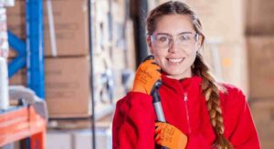 medium closeup portrait of a smiling young woman in a warehouse setting with industrial shelves and boxes. The woman wears safety glasses, red jacket and orange gloves, and holds a mop by its handle.