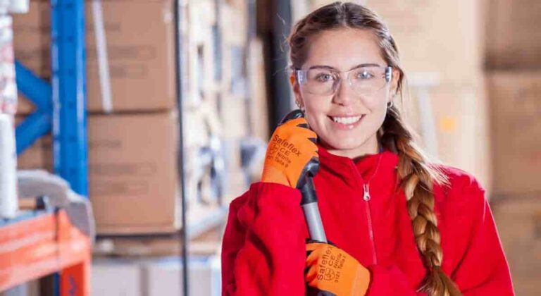 medium closeup portrait of a smiling young woman in a warehouse setting with industrial shelves and boxes. The woman wears safety glasses, red jacket and orange gloves, and holds a mop by its handle.