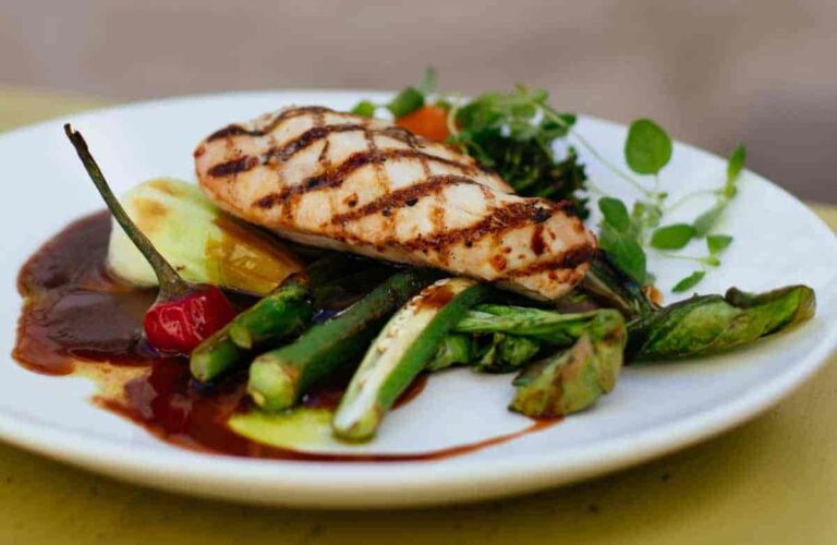 Grilled chicken breast with char marks sits atop mixed vegetables and drizzle of sauce, garnished with fresh greens, all on a white plate.