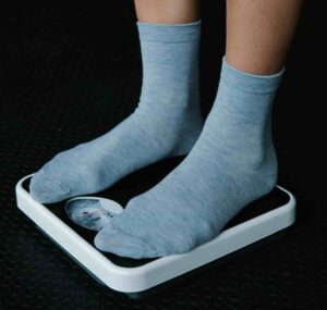 Close-up of a person in blue-gray socks standing on a white analog weighing scale against a dark floor. The scale's needle points to an indistinct measurement.