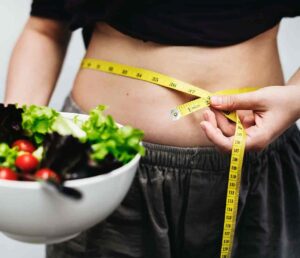 A woman's bare stomach with a crop top and black shorts. The woman is holding a bowl of salad and in her other hand is holding a tape measure wrapped around her waist.