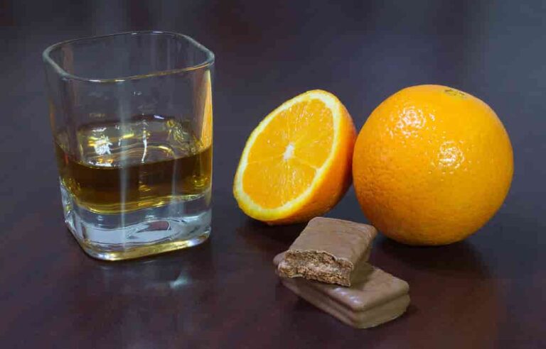 A glass of whiskey is next to a whole orange and an orange half. Two chocolate bars are stacked nearby on a dark surface