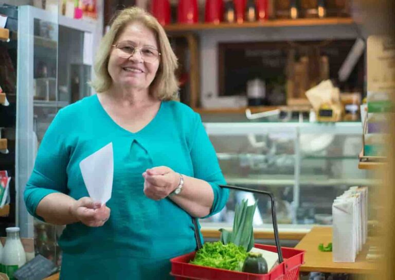 Smiling woman in a teal shirt holds a shopping list in a grocery store. A red basket with vegetables is on the counter